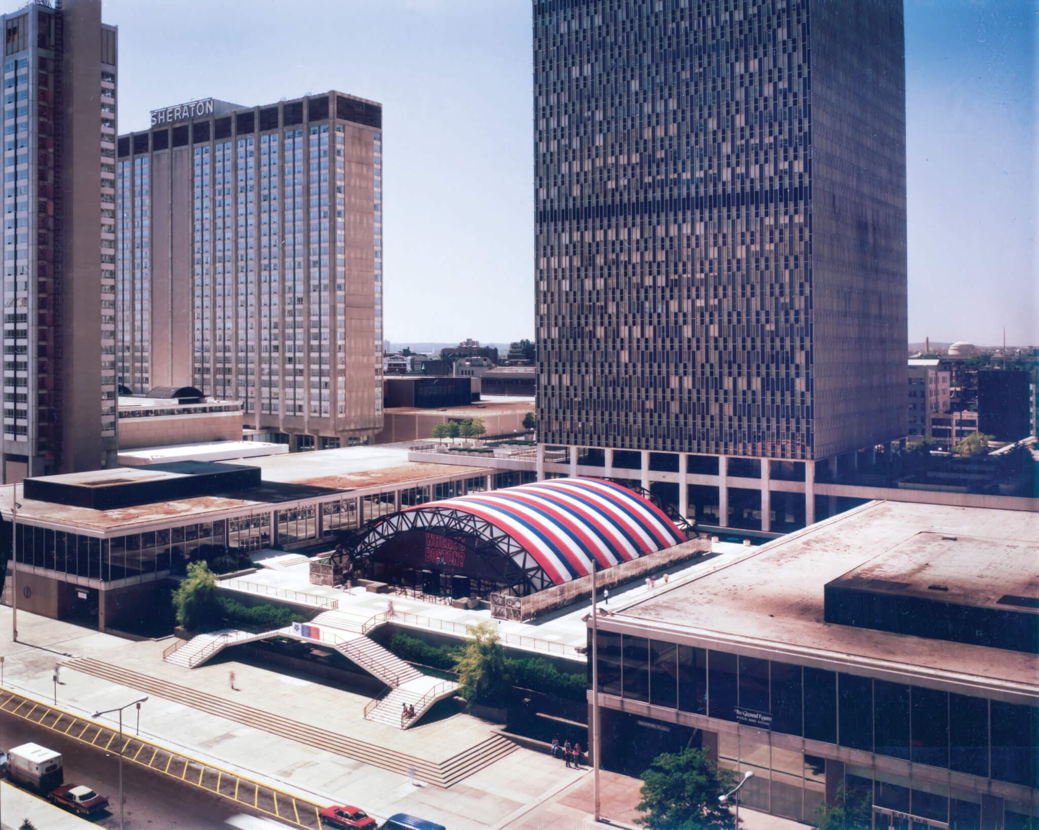 A zoomed out image of a red, white, and blue pavilion between city buildings.
