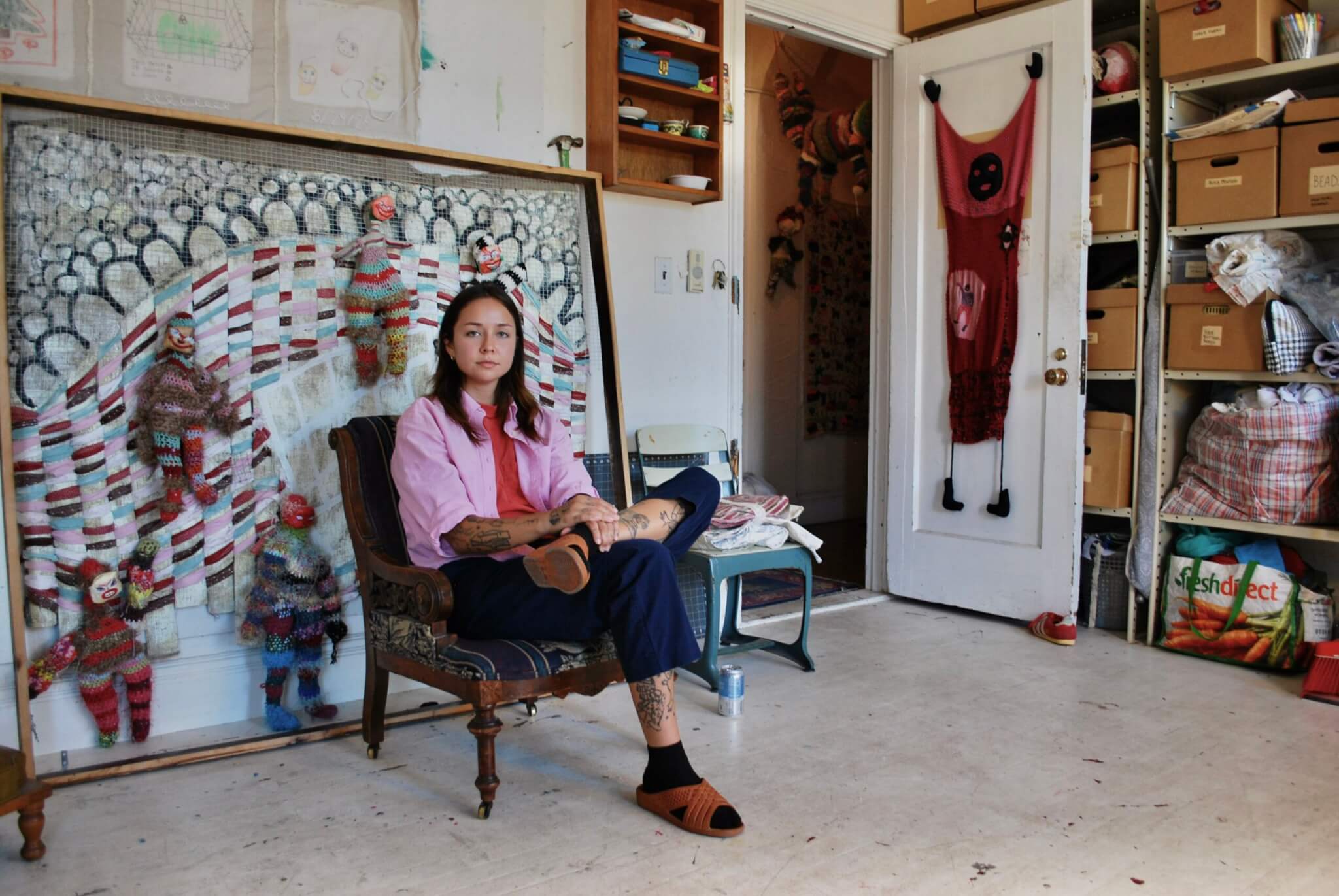A woman is seated in an armchair in a studio featuring crocheted artworks.