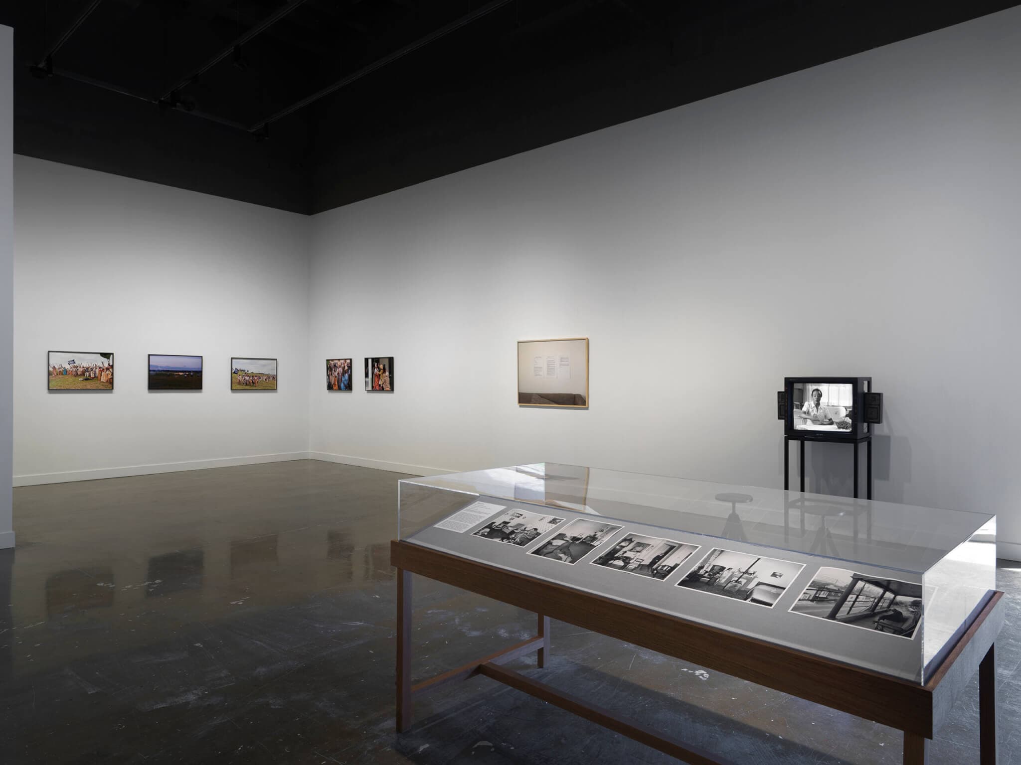 In the foreground on the right, a freestanding wooden vitrine is filled with grayscale photographs: these include a figure sprawled on a chair and a man sitting limply on a porch. Other artworks fill the background, including a boxy monitor depicting a seated woman and a framed photograph of printed pages of text hanging on a wall.